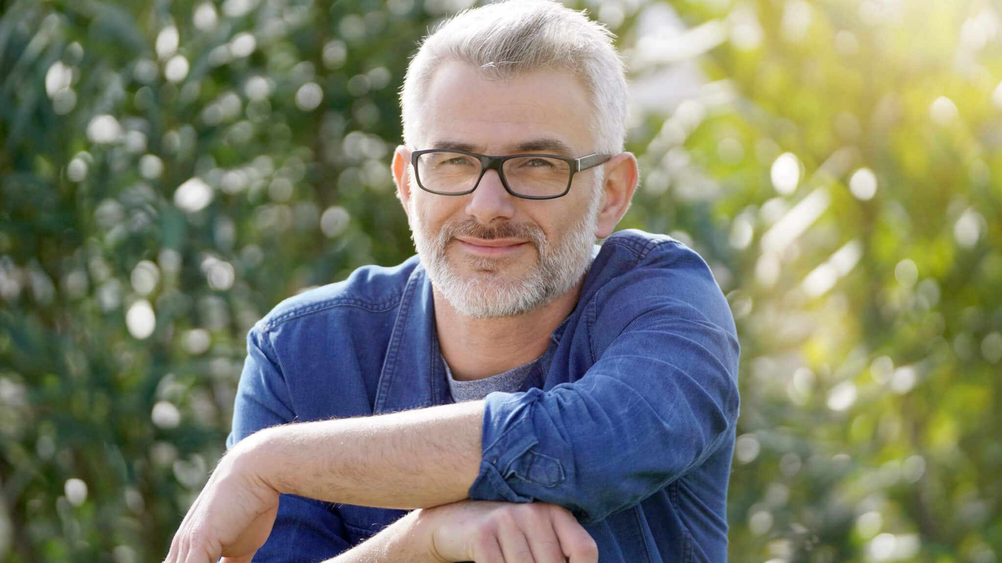 Smiling, contemplative man in his fifties wearing a blue plaid flannel, thinking about his debt-free future on a sunny day outdoors.