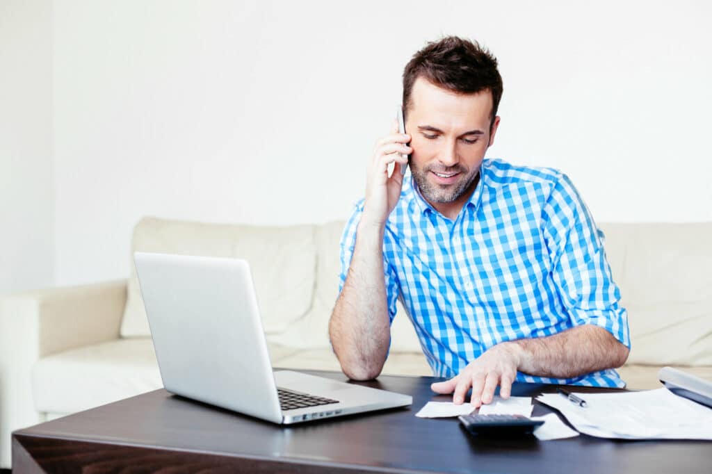 Man in blue plaid shirt working on a calculator and papers on laptop