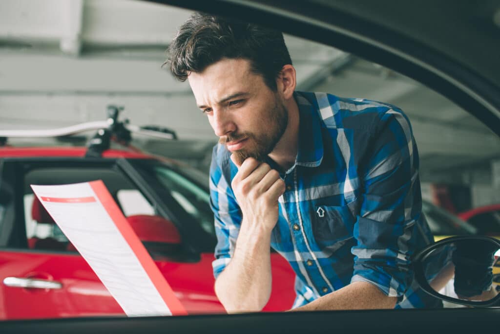 A young man sitting at a desk, looking at paperwork, with a new red vehicle parked in the background.