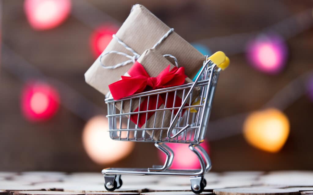 Holiday shopping portrayed by cart, holiday lights and gift in cart