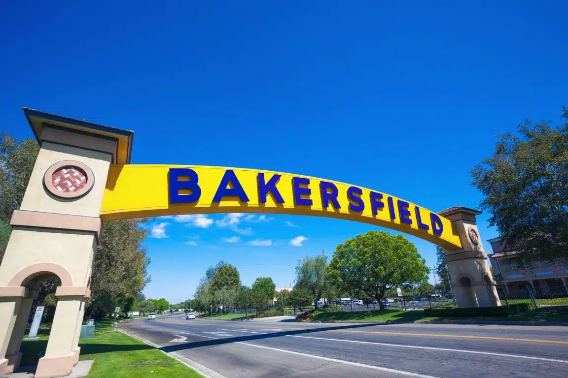 Iconic yellow Bakersfield sign arching over a roadway with blue sky and trees in the background.