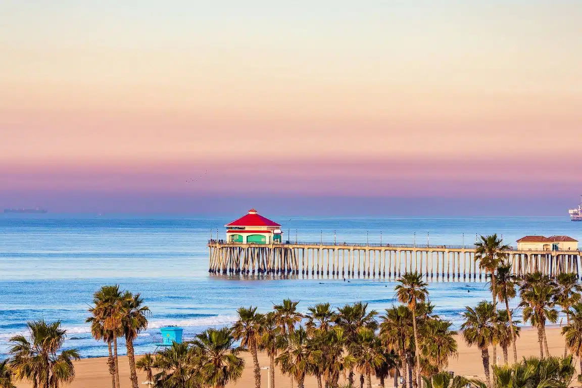 Huntington Beach Pier at sunset with red-roofed pavilion and palm trees along the sandy shoreline.