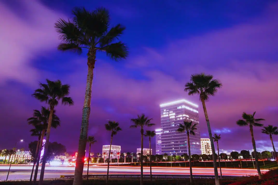 Night view of Irvine business district with palm trees, light trails, and a glass office tower under a purple sky.