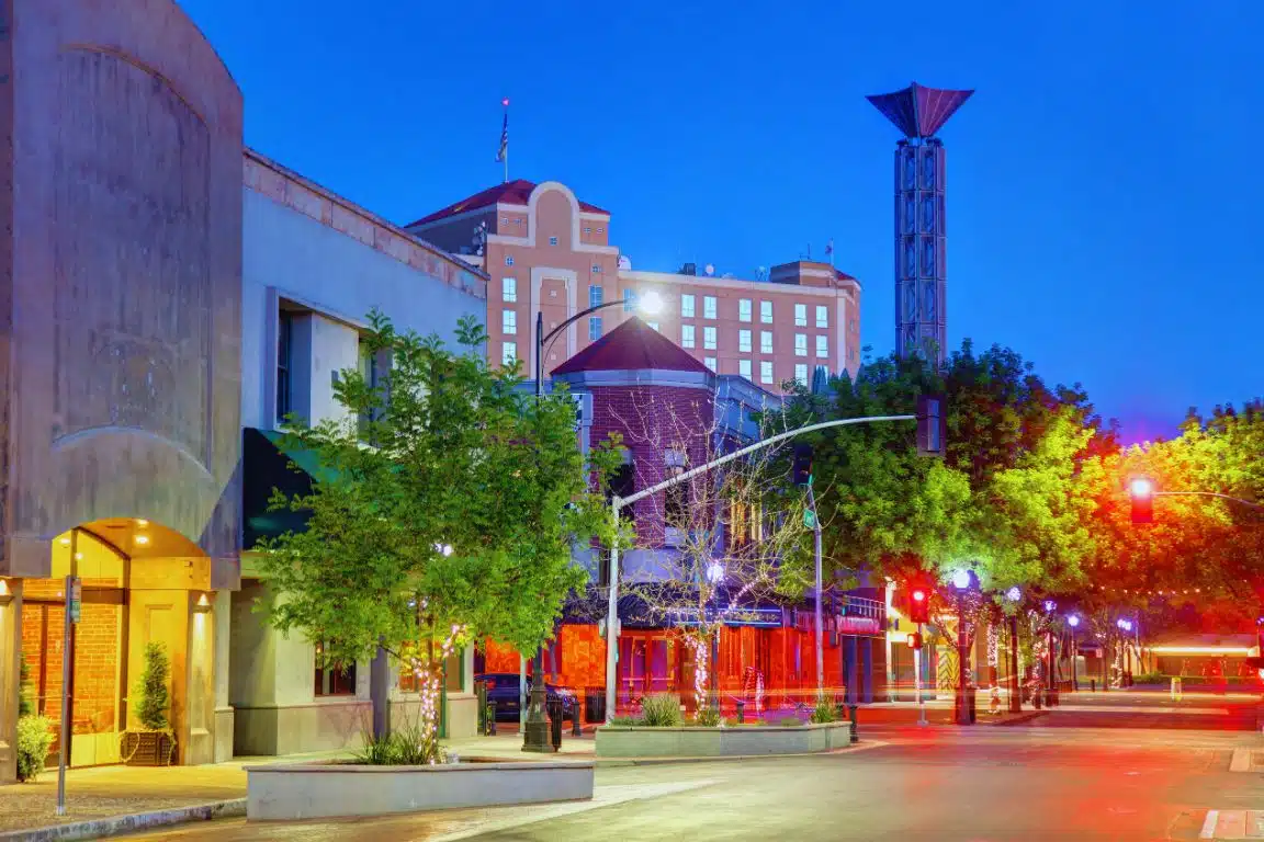 Downtown Modesto at dusk with streetlights, trees, and historic buildings along a quiet city street.