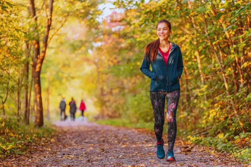Smiling woman walking on a leafy wooded trail in autumn, with three distant walkers on the path.