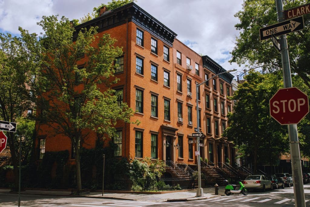 Brooklyn brownstone rowhouses on a tree-lined street with stoops, corner stop signs, and a Clark St one-way sign.