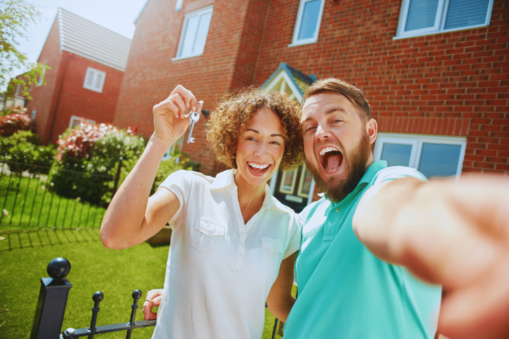 Smiling couple taking a selfie in front of a brick house, celebrating a new home on a sunny day.