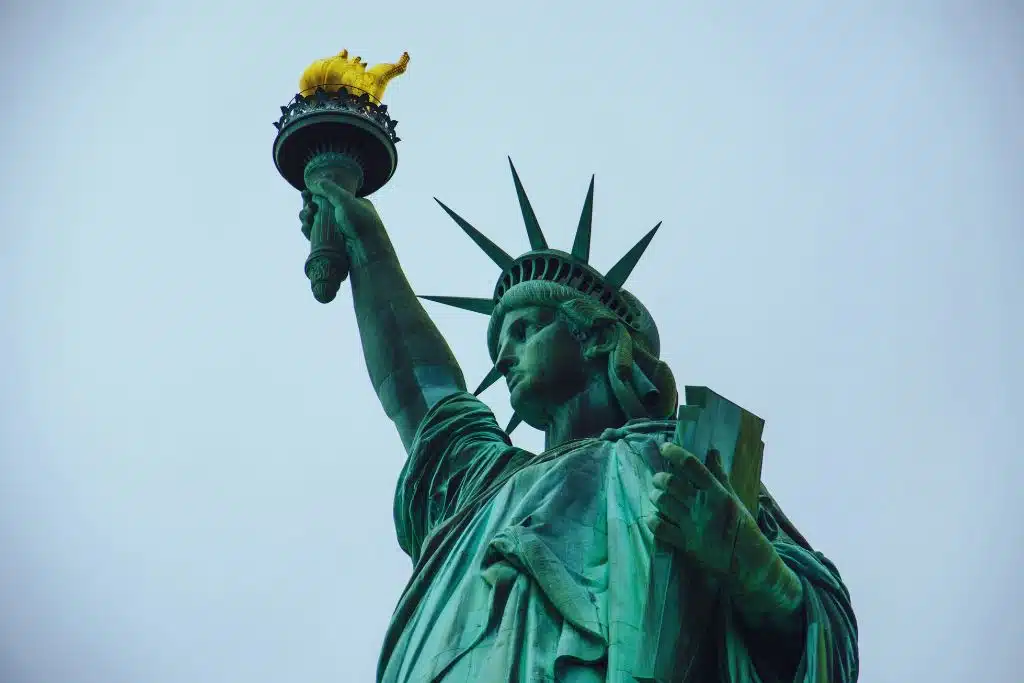 A close view of the Statue of Liberty in New York Harbor, highlighting the raised torch, spiked crown, and verdigris copper surface against a soft sky.