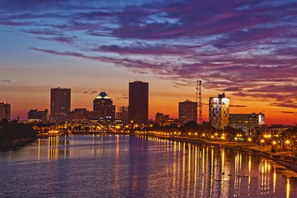 Rochester skyline at dusk with High Falls waterfall illuminated and city lights reflecting on the Genesee River.