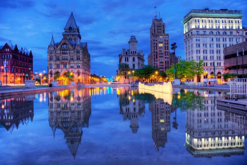 Syracuse skyline at dusk with illuminated buildings and Clinton Square fountain reflecting city lights.