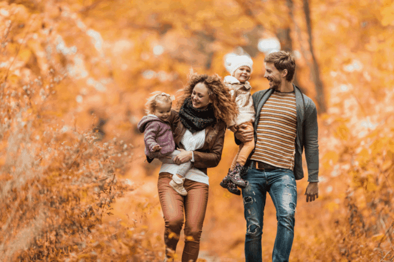 Family walking together on a tree-lined path with colorful fall leaves, relaxed and smiling in cool weather.