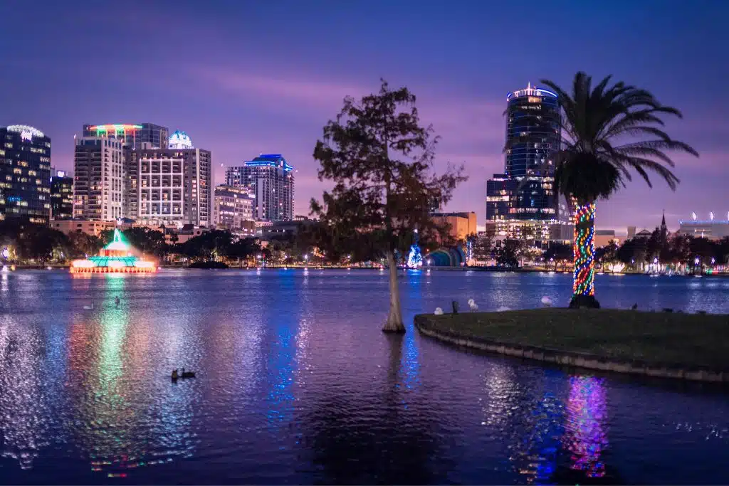 Orlando skyline at night with illuminated buildings, lake reflections, and theme park fireworks in the distance.